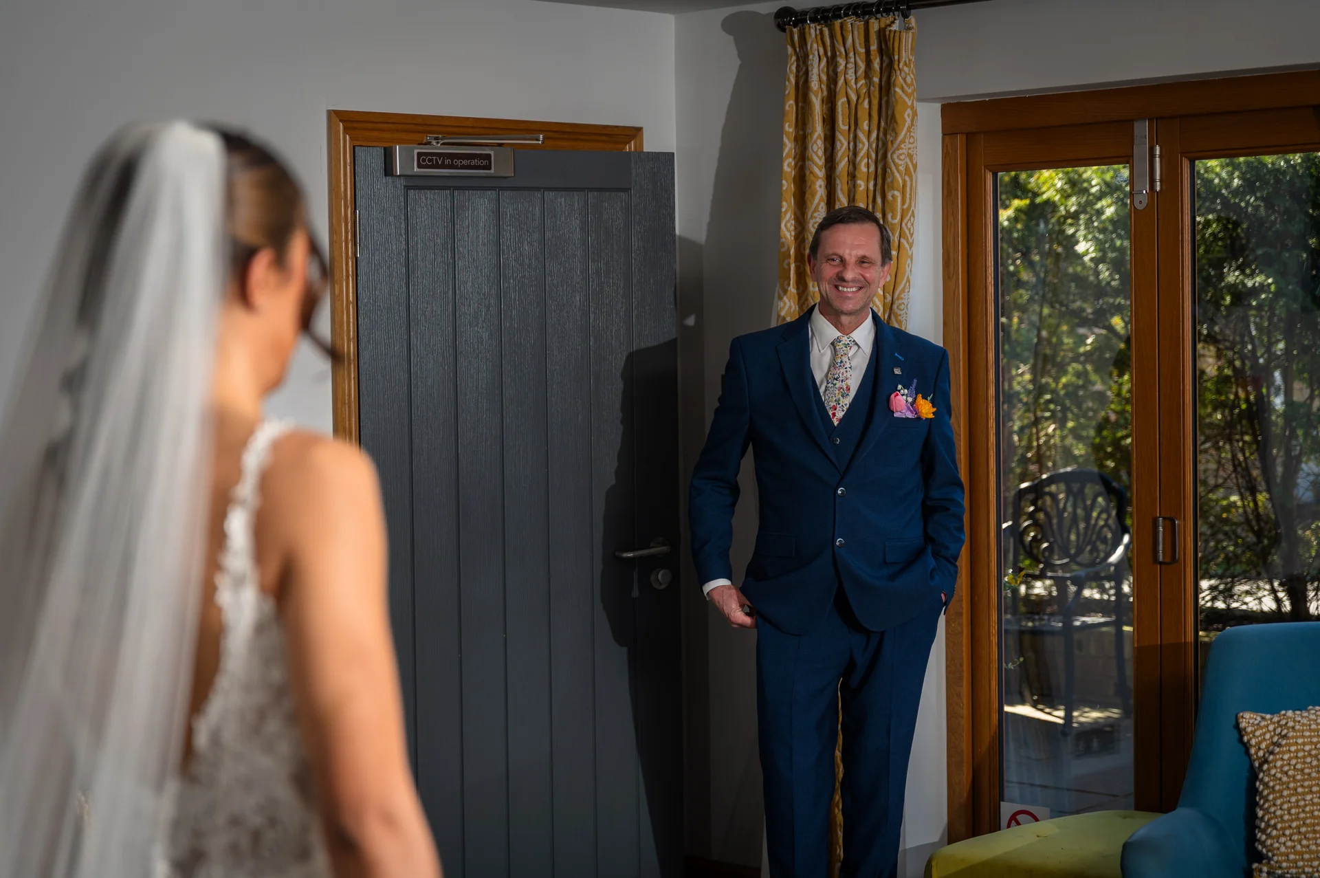 A smiling father in a blue three piece suit proudly looks on during a first look moment with his daughter wearing a wedding dress and veil.