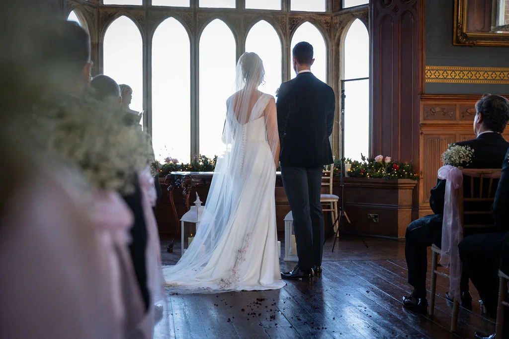 A bride and groom stand facing a large arched window during their wedding ceremony inside a grand stately home with wooden floors.