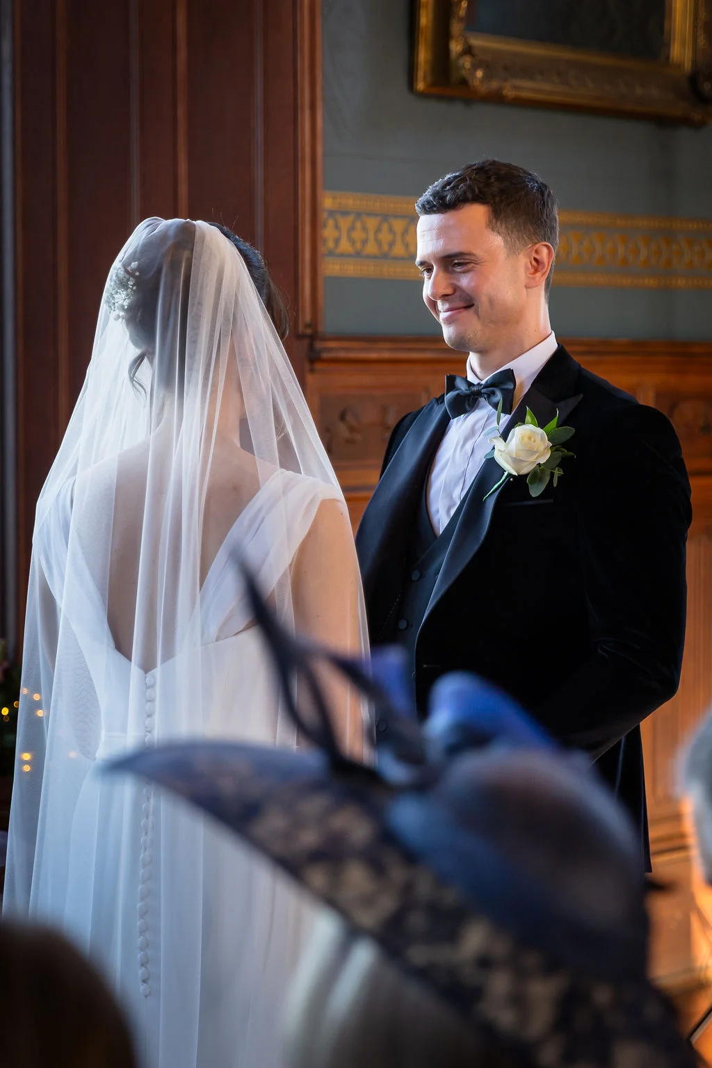 A groom looks lovingly at his bride during the ceremony inside a wood-panelled country house room, wearing a classic black tuxedo while she wears a simple white gown with a veil.