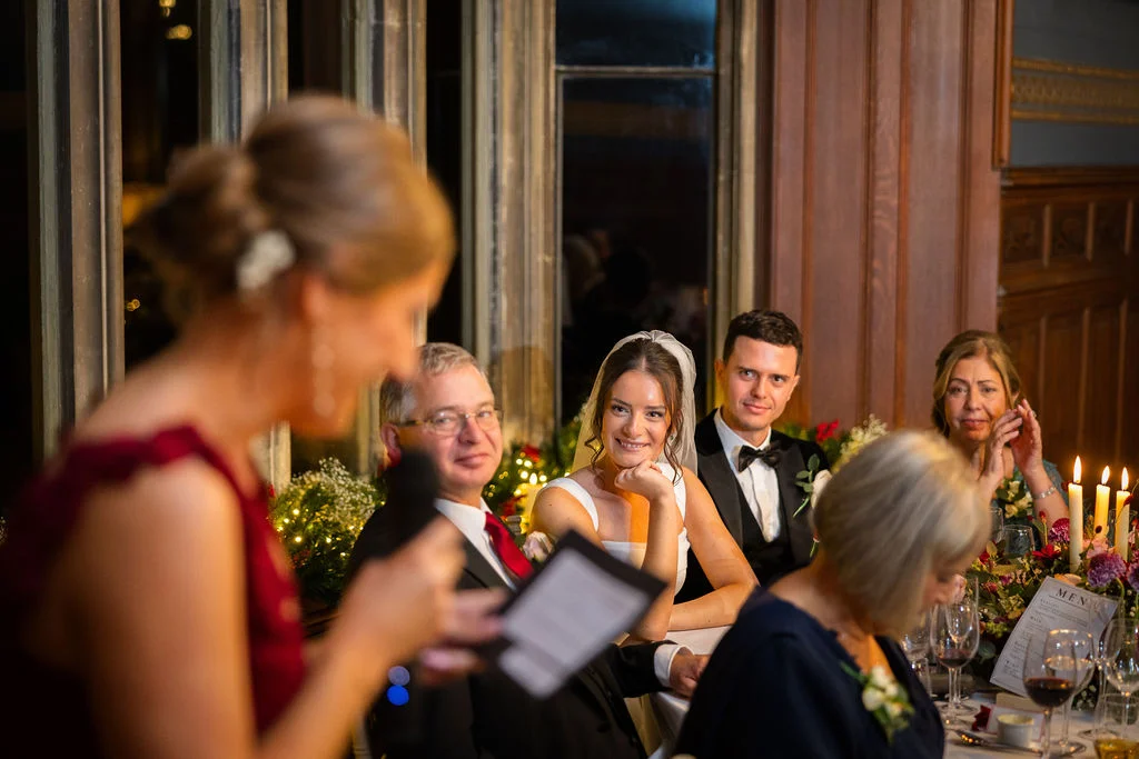 A smiling bride and groom listen to a guest giving a speech at the top table, illuminated by warm candlelight in a wood-panelled reception room.