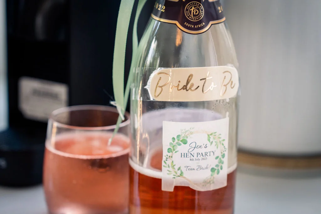 A close-up shot shows a personalised bottle of sparkling wine featuring a custom 'Bride to Be' label and a botanical hen party sticker, paired with a glass of pink drink.