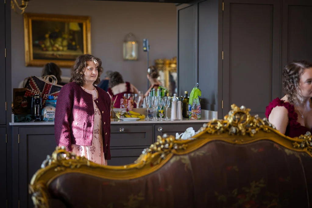 A candid shot capturing wedding guests getting ready inside a room featuring dark grey cabinetry, ornate gold-framed furniture, and natural window light.