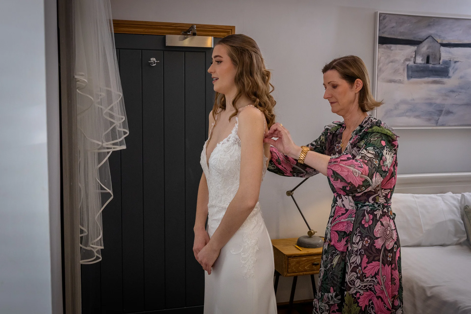 The mother of the bride carefully fastens her daughter's elegant lace and crepe wedding dress during morning preparations.