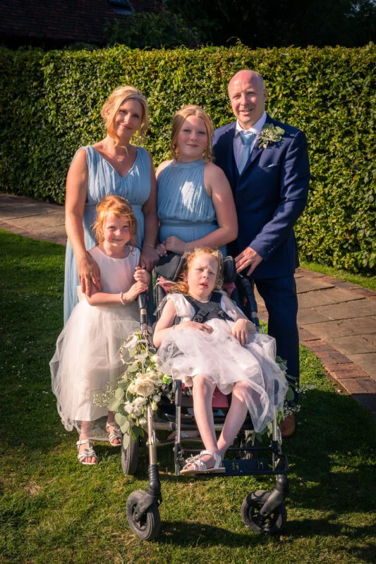 A happy family posing together outdoors in a sunny garden setting, with a groom in a navy suit, women in blue dresses, and children in white flower girl dresses.