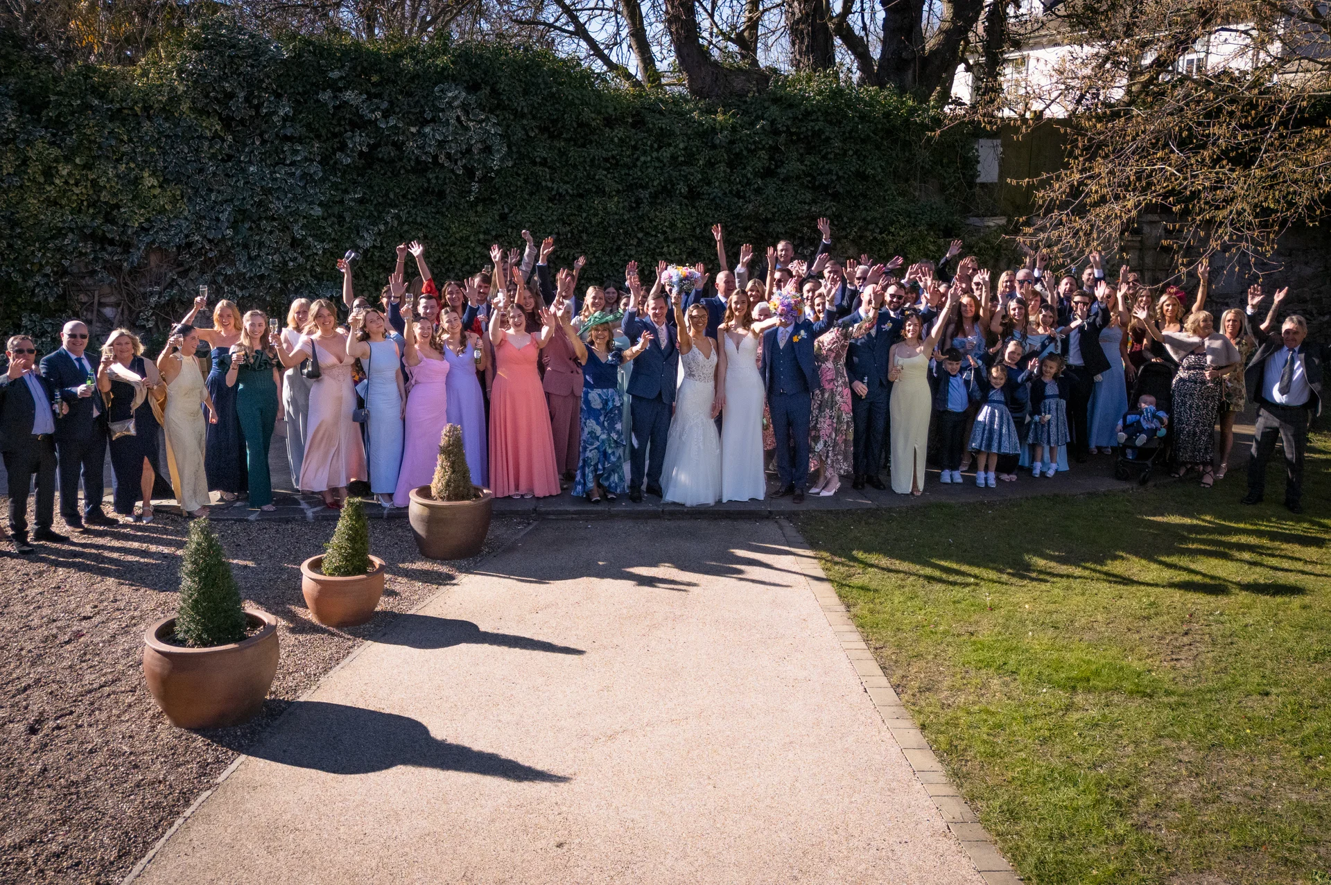 A large group of wedding guests stand outdoors in a sunny garden setting, waving their hands in the air with joyful expressions.