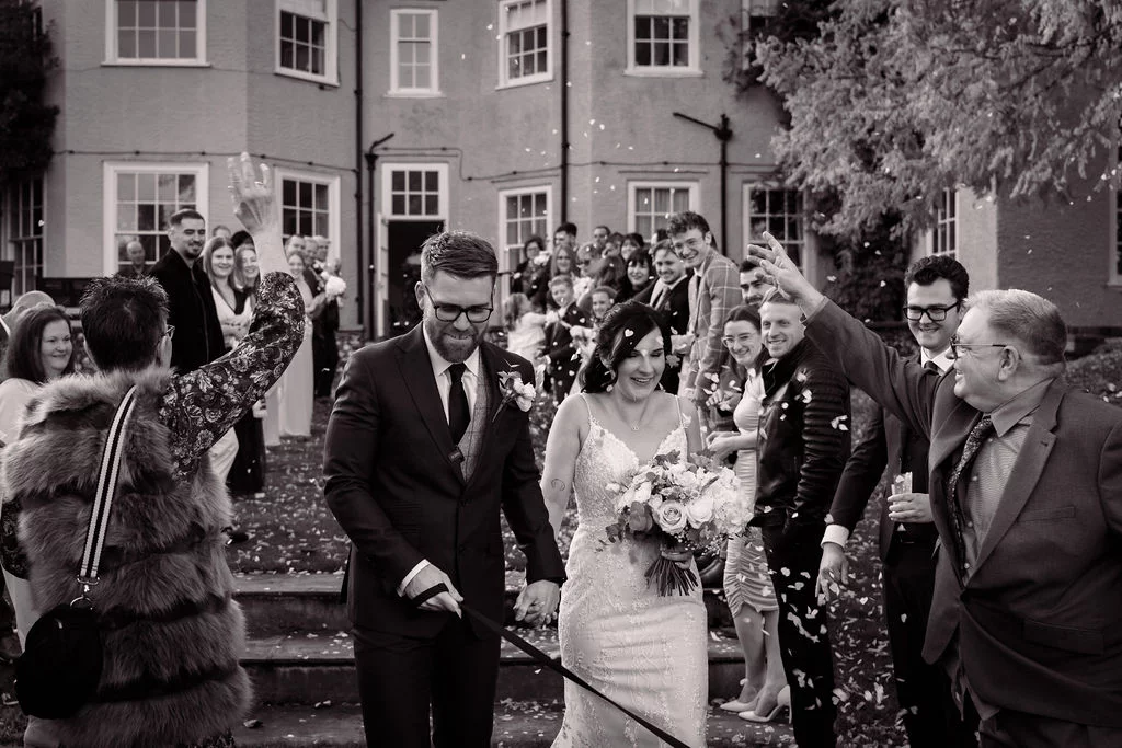 A joyful newlywed couple walks hand in hand down the stone steps of a country house as smiling wedding guests shower them in confetti.