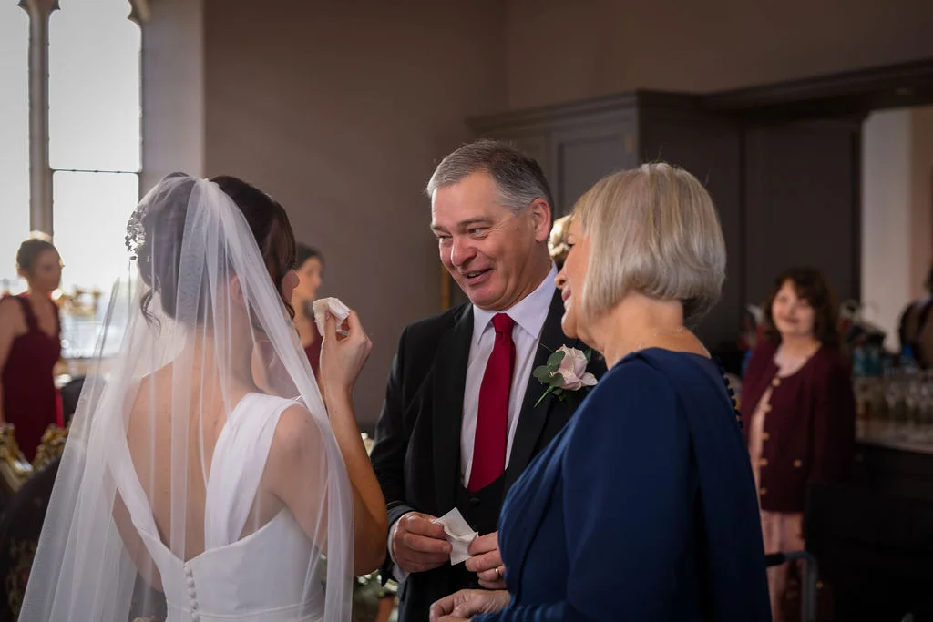 A smiling father of the bride wearing a red tie looks affectionately at the bride while wiping a tear, alongside the mother of the bride indoors.