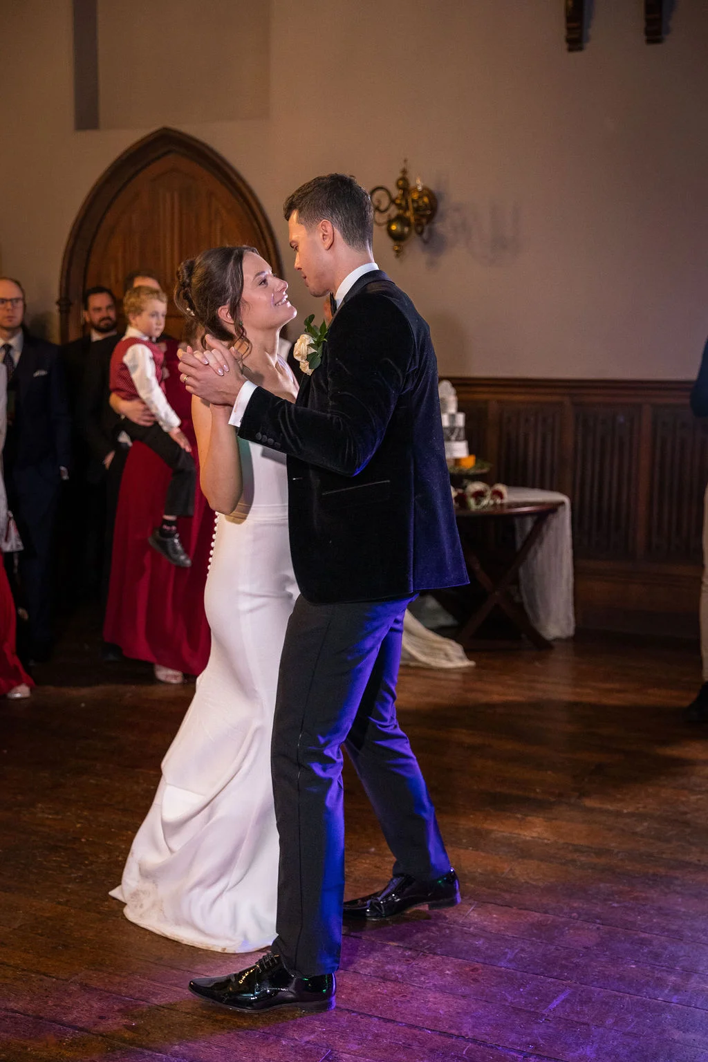 Newlyweds share their first dance together on a wooden floor inside a historic hall with wood panelled walls and watching guests.