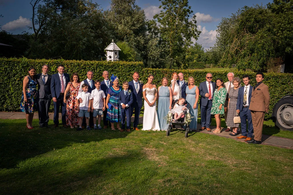 A large wedding party and family group pose together outdoors on a sunlit lawn against a tall green hedge.