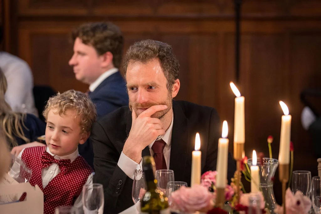 A guest and a young boy wearing a bow tie sit attentively at a wedding breakfast table illuminated by candlelight during speeches.