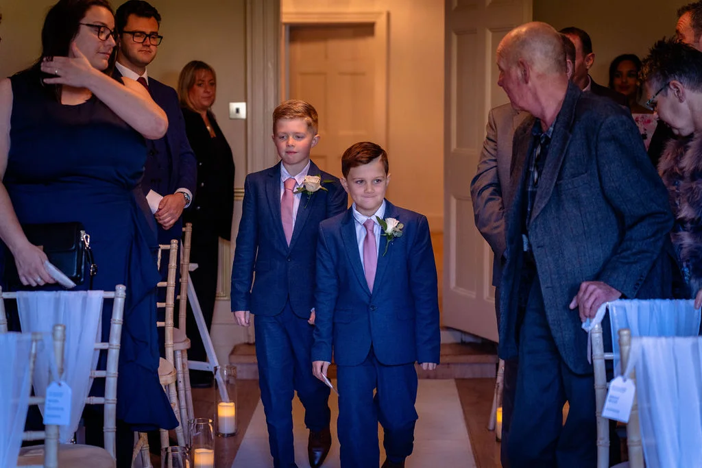 Two page boys in matching blue suits and pink ties walk down the aisle while guests look on during a wedding ceremony.