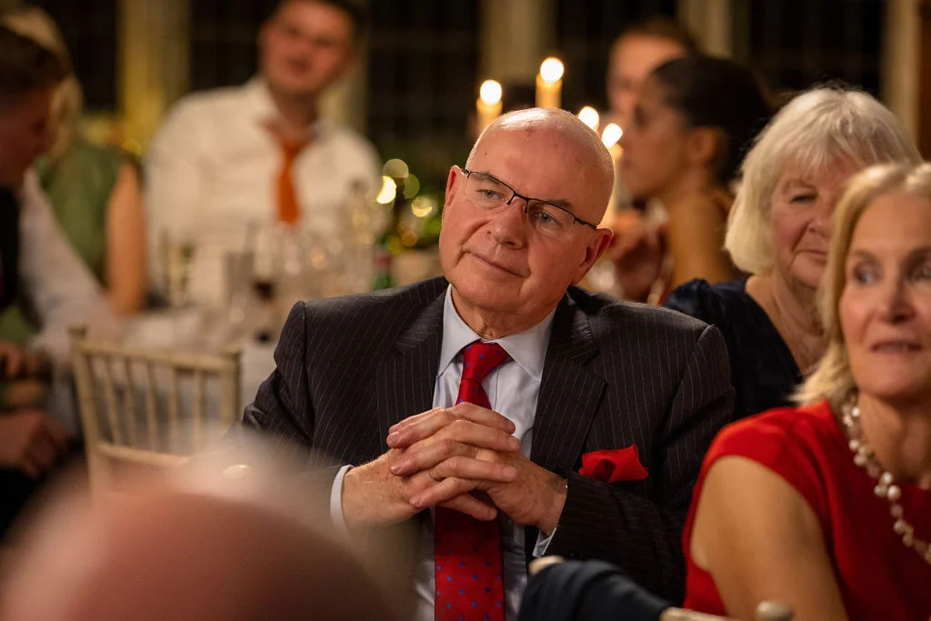 An older male guest in a pinstripe suit and red tie listens attentively with clasped hands during the wedding reception speeches.