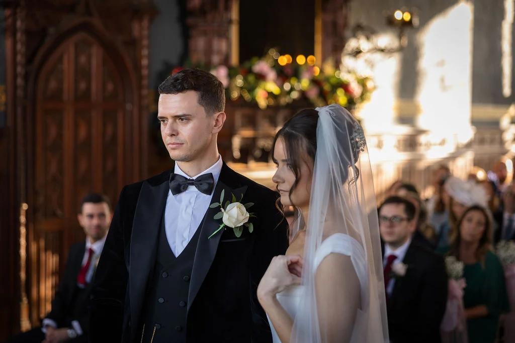 A newlywed couple stands together during their church ceremony, with the groom in a classic black velvet tuxedo and the bride in a simple white gown and veil.