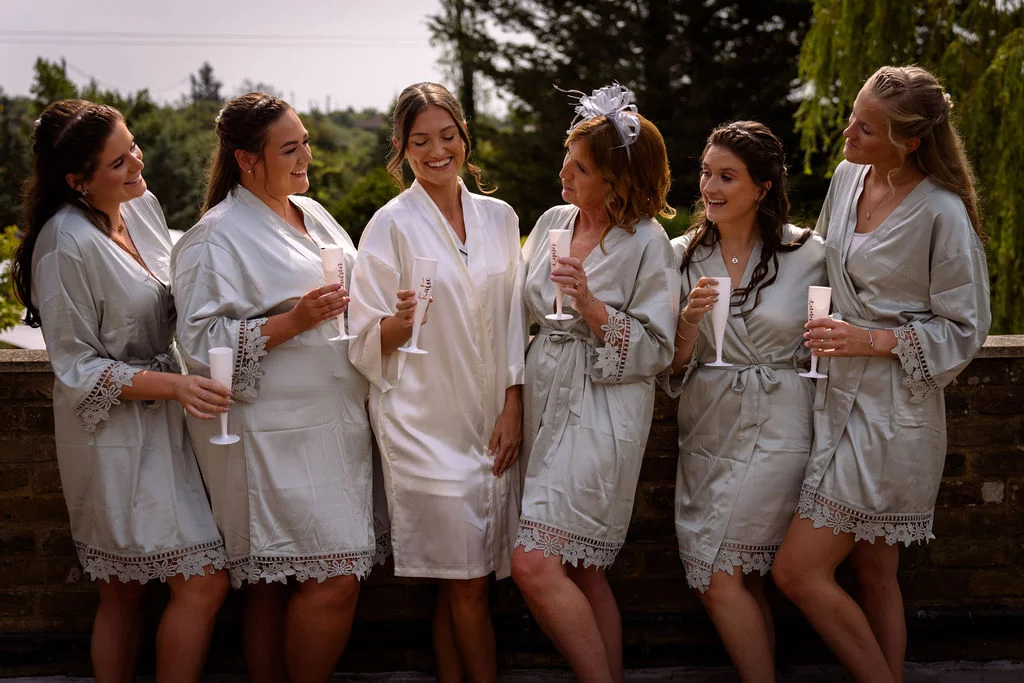 The bride in a white robe and her bridal party in matching sage green satin robes stand outdoors laughing and holding champagne flutes.