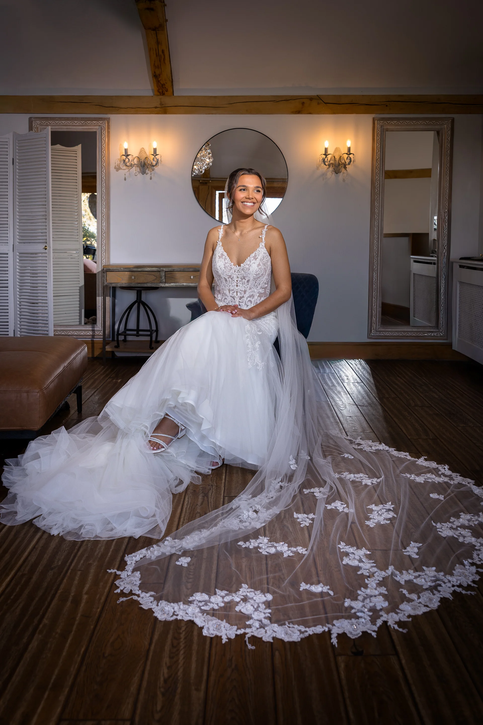A smiling bride sits gracefully in a rustic barn venue, wearing an A-line lace wedding dress with a dramatic cathedral-length veil spread across the wooden floor.