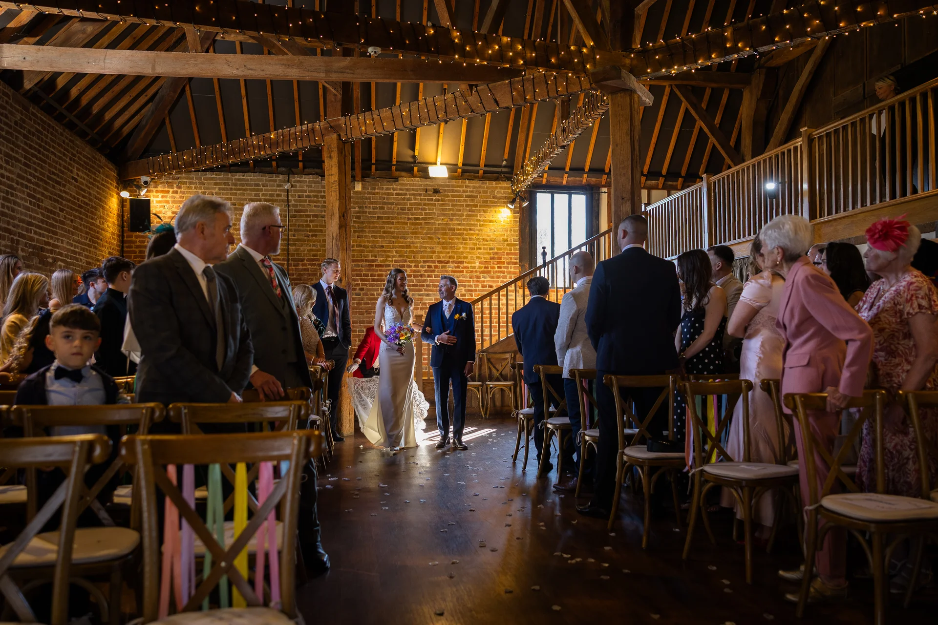 A bride walks down the aisle with her father inside a rustic barn venue decorated with exposed wooden beams and a glowing fairy light canopy.