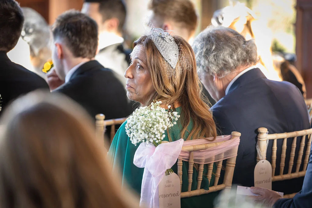 A wedding guest wearing a silver fascinator and green dress looks back while seated in a row of chiavari chairs decorated with reserved signs and floral pew ends.