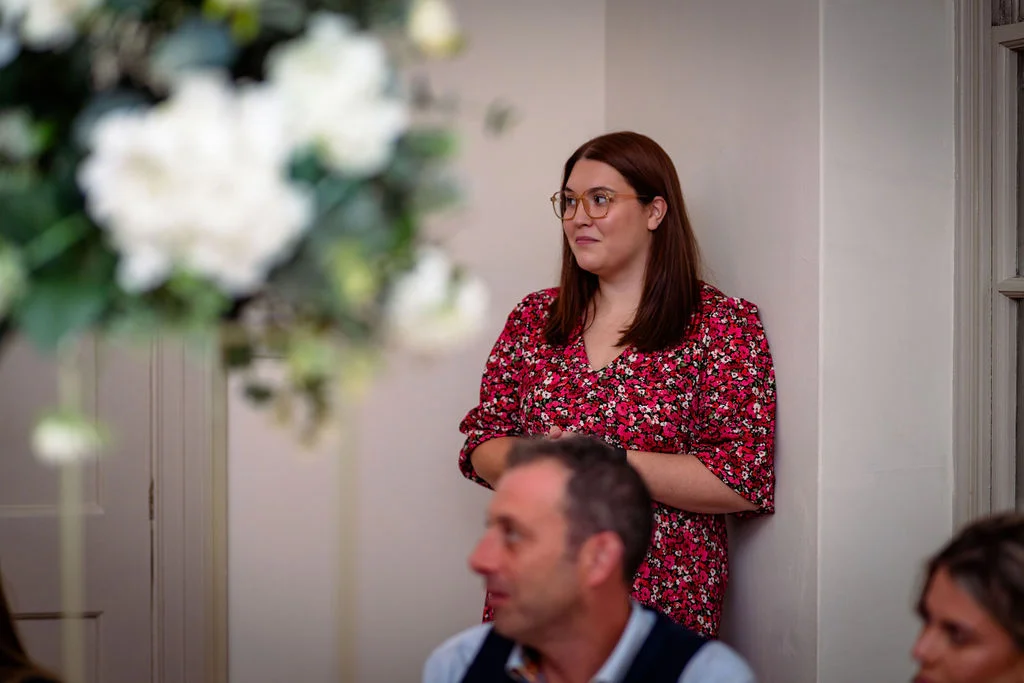 A wedding guest wearing a red floral print dress and glasses looks on with a soft smile during the wedding speeches, framed by white floral arrangements in the foreground.