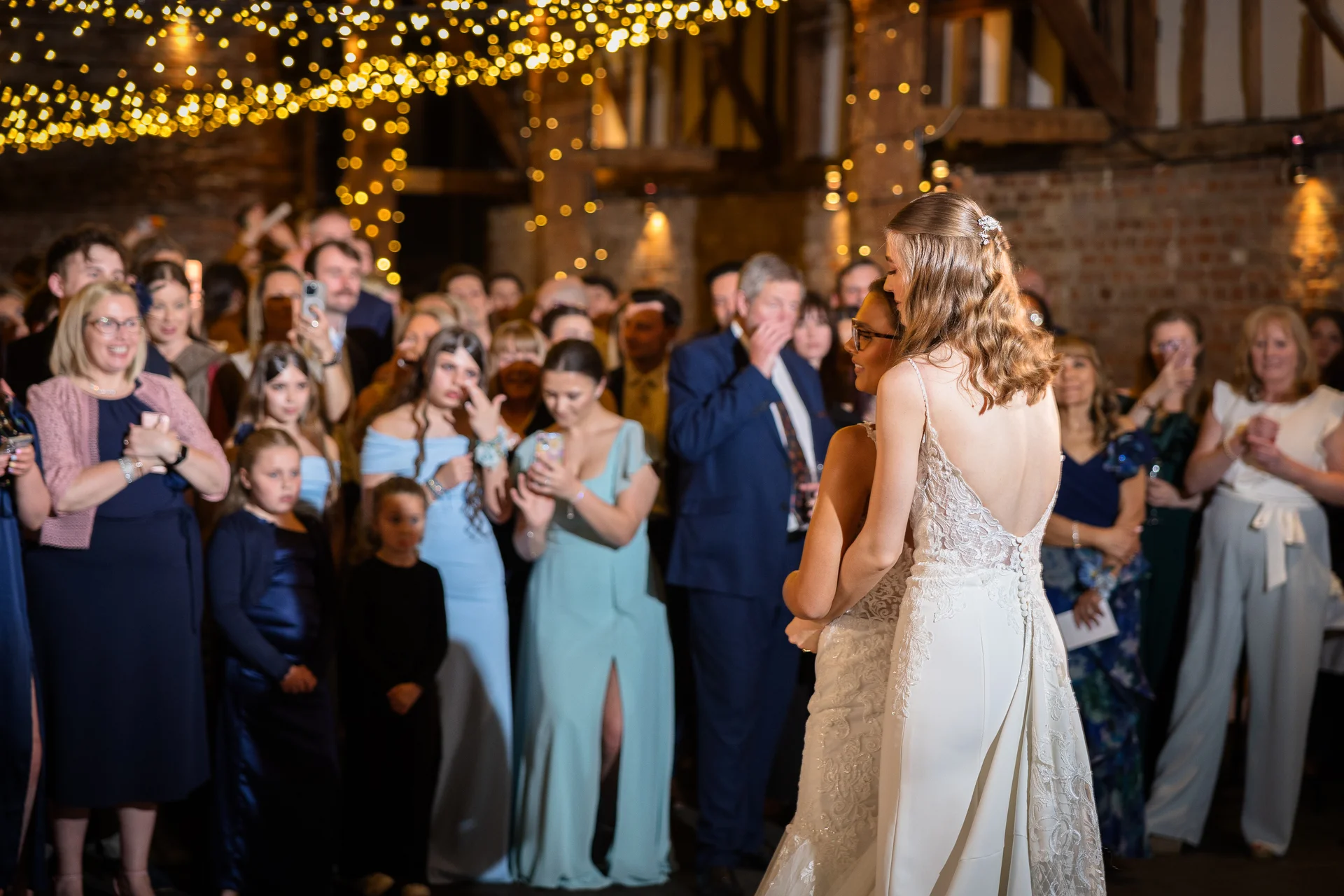Two brides embrace during their first dance inside a rustic barn venue illuminated by a warm fairy light canopy while guests watch.