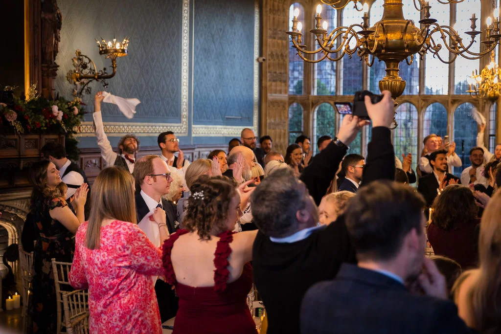 Wedding guests in a grand historic hall wave their napkins in the air and cheer excitedly during the speeches.