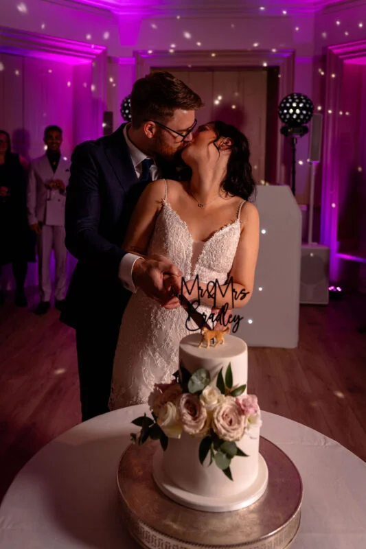 A bride and groom share a tender kiss while cutting their elegant two-tiered white wedding cake adorned with fresh roses under vibrant purple reception lighting.
