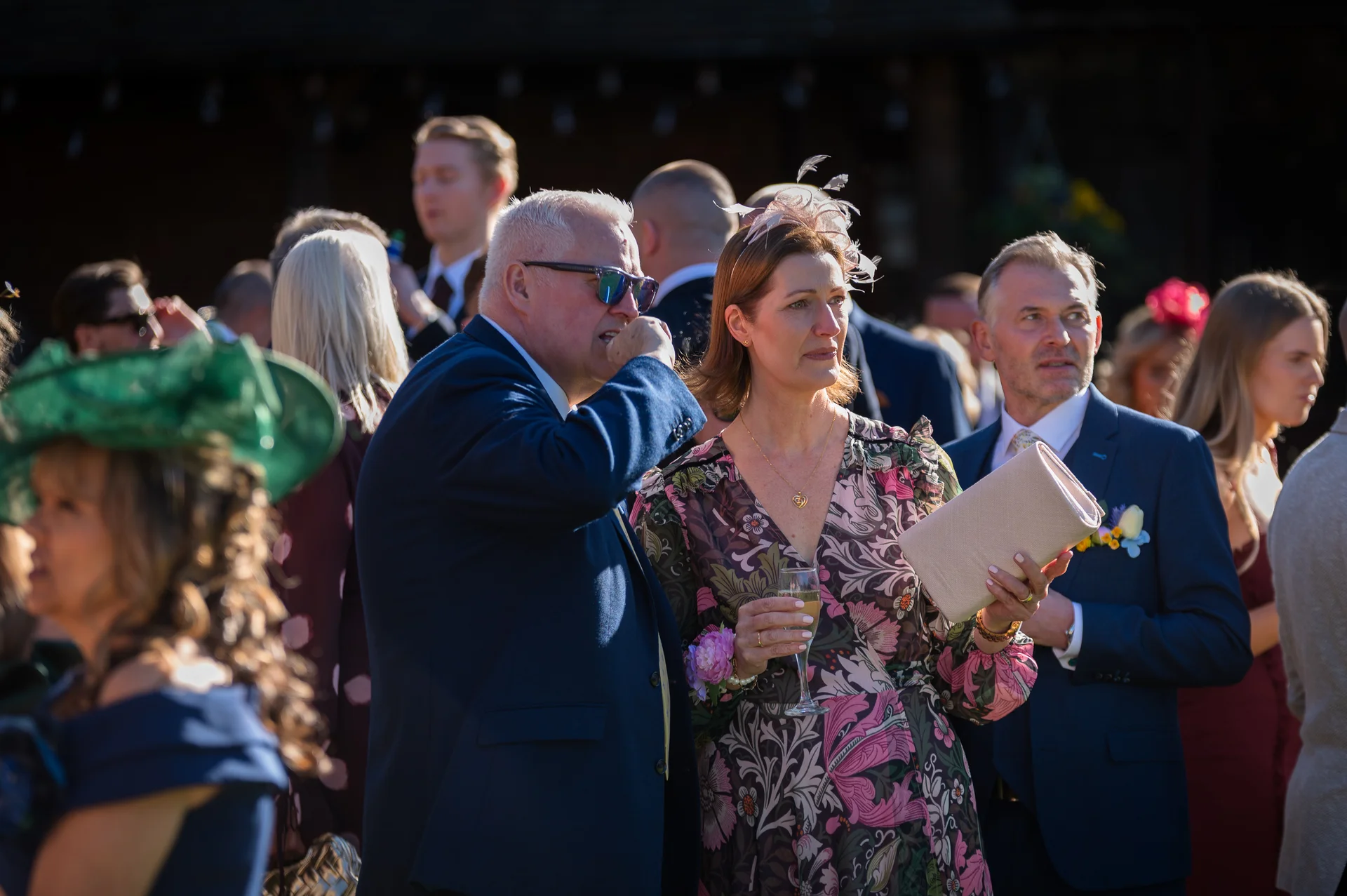 Wedding guests mingle outdoors during a sunny drinks reception, bathed in warm golden hour light.