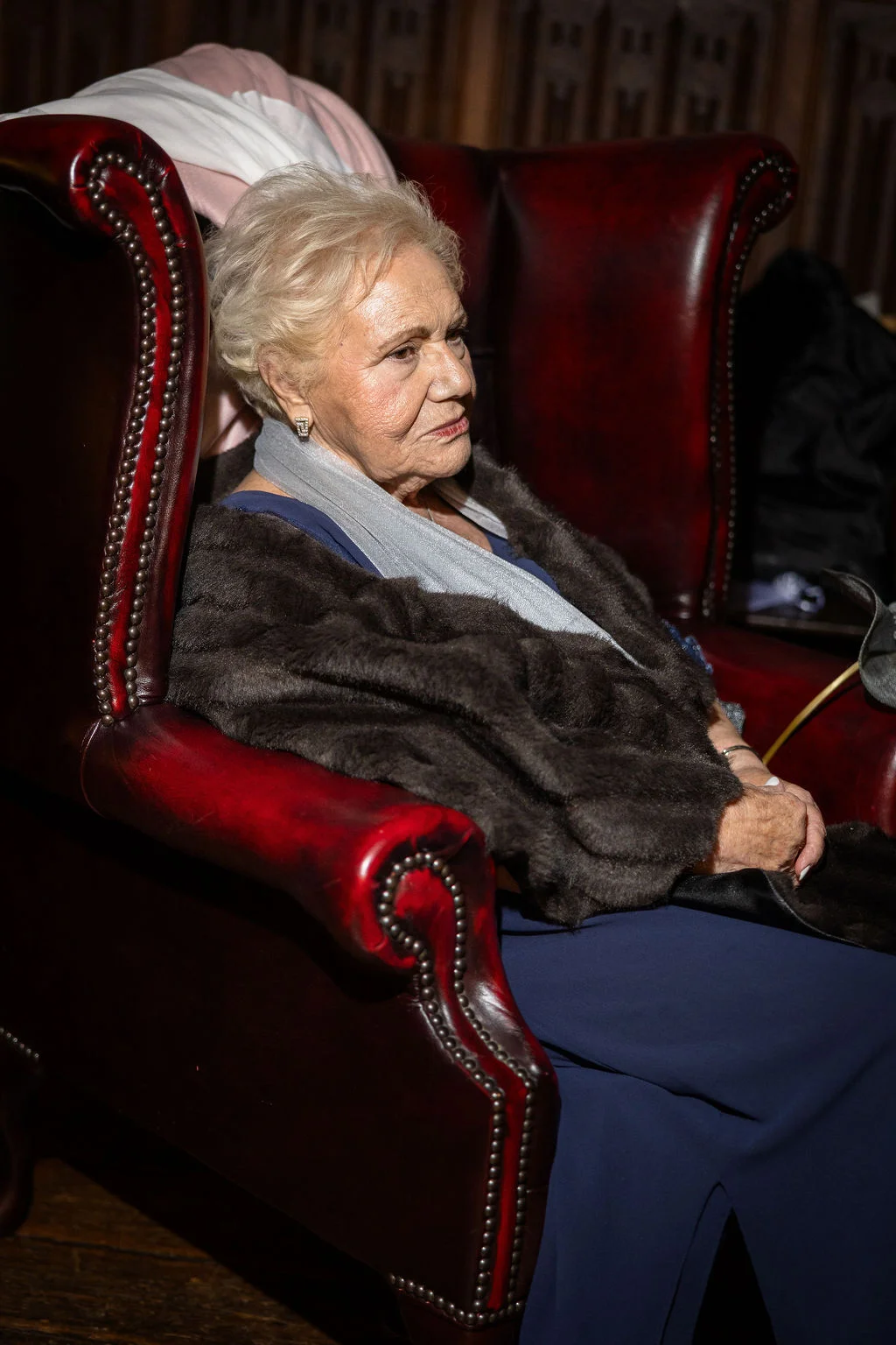 An elderly female guest relaxes in a deep red leather armchair, wearing an elegant blue outfit and a dark fur wrap.