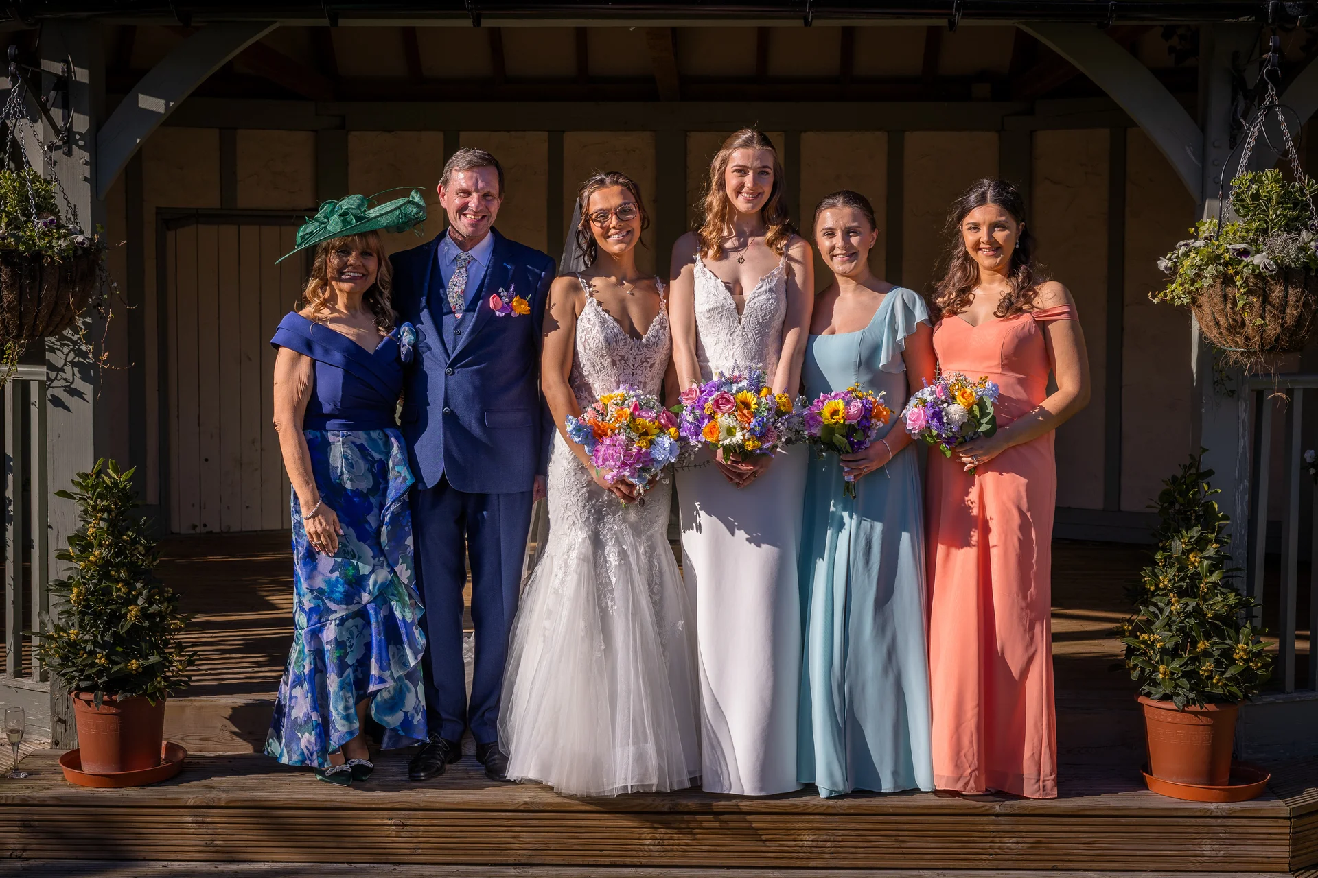 A joyful wedding group including parents, two brides, and bridesmaids holding colourful bouquets poses together on a wooden terrace under bright sunlight.