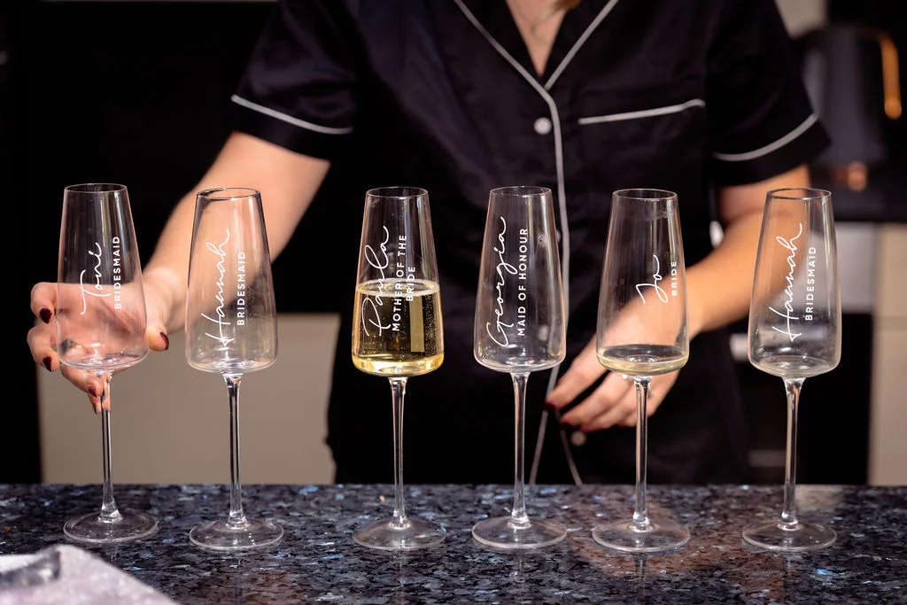 A person wearing black pyjamas with white piping arranges a line of six personalised champagne flutes featuring names and wedding roles on a dark countertop.
