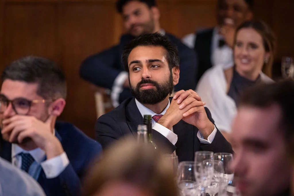 A guest sits at a wedding reception table, attentively watching the speeches with hands clasped together while other guests laugh in the background.