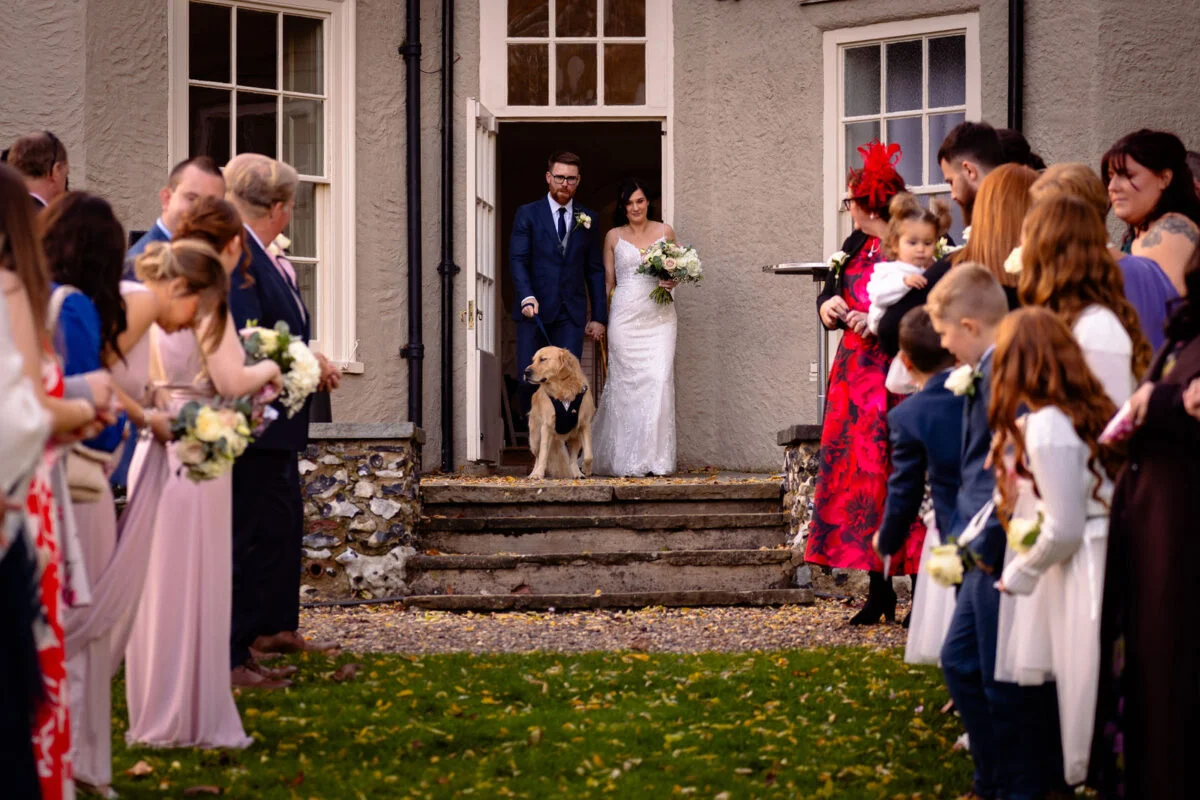 The bride and groom step out of a country house doorway holding hands alongside their golden retriever while wedding guests cheer from either side of the path.