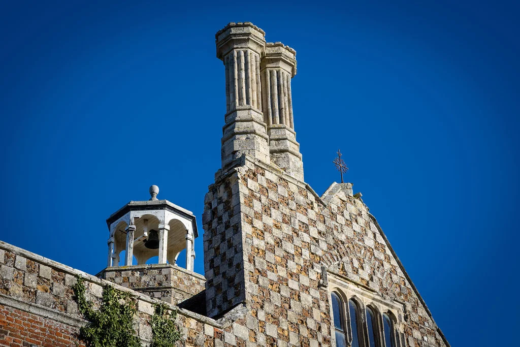 An exterior view of a historic country house featuring detailed stone chequerwork, twin chimney stacks, and a small bell turret set against a clear blue sky.