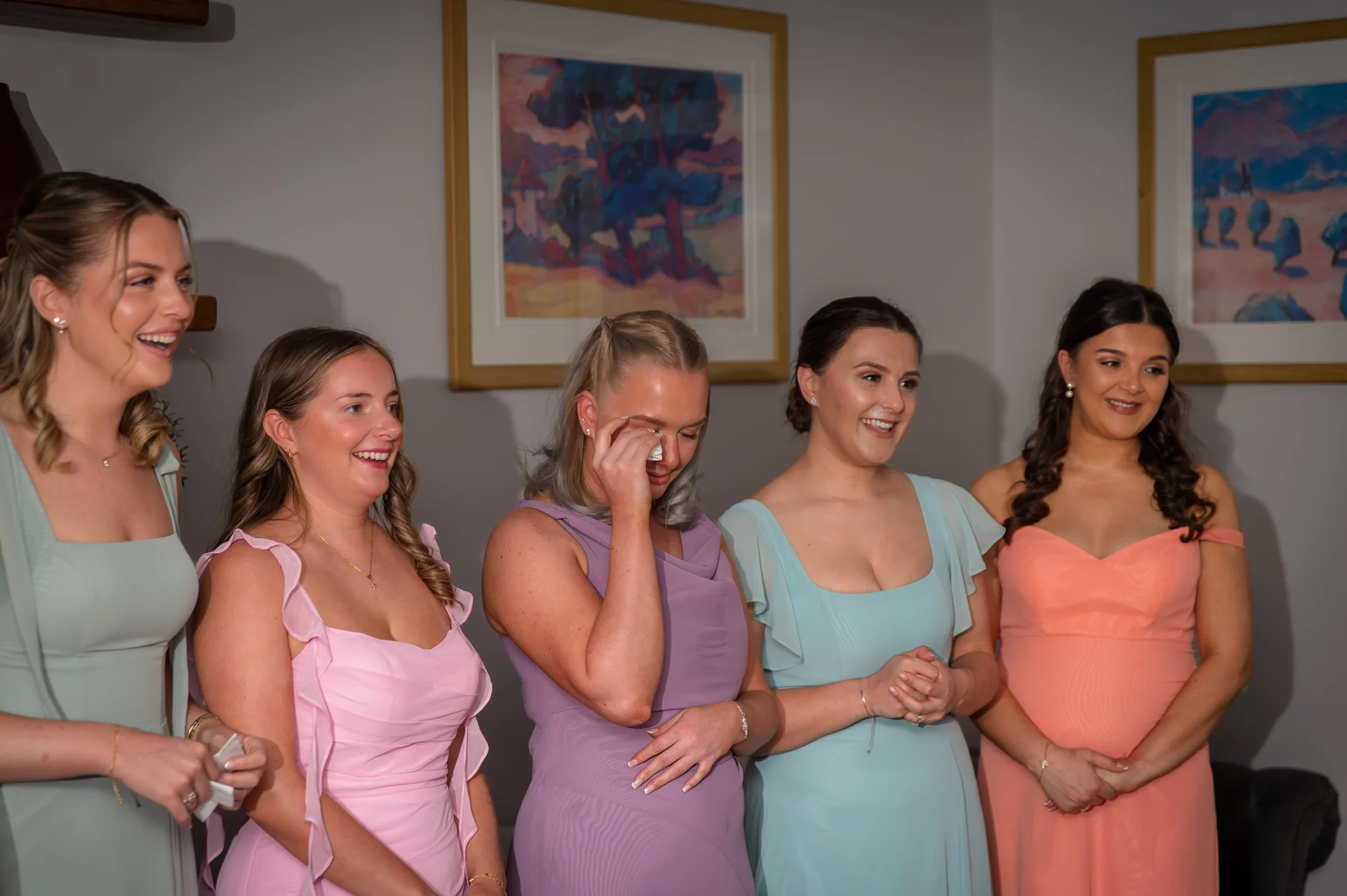 A group of five bridesmaids in colourful dresses stand together indoors, with one wiping away happy tears while the others look on with emotional smiles.