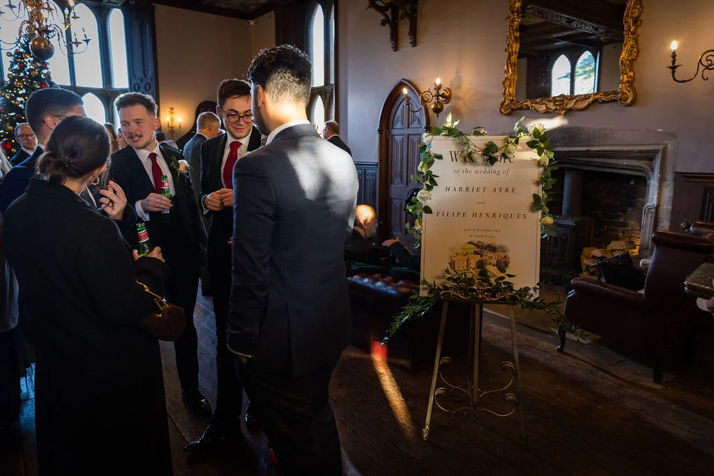 Guests mingle in a historic manor house next to an elegant welcome sign adorned with foliage during a winter wedding reception.