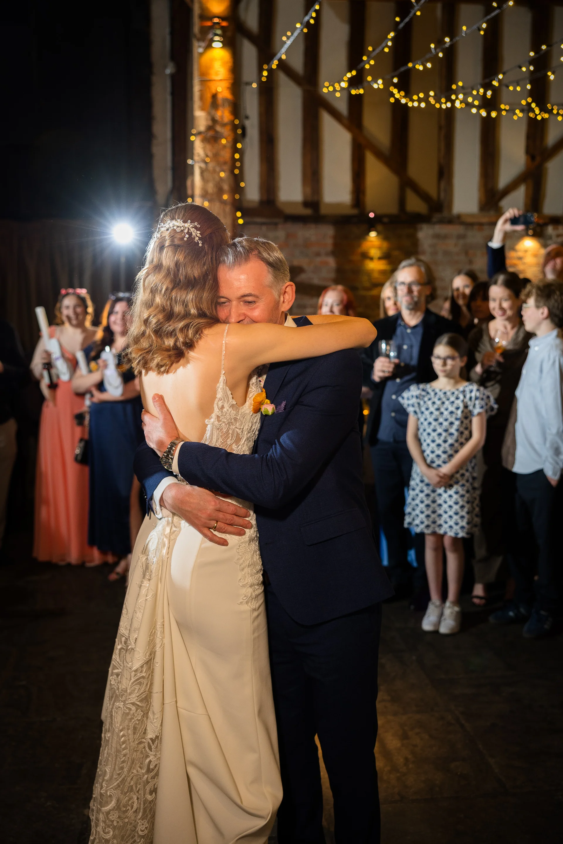 A bride and her father share an emotional embrace during their dance inside a rustic barn venue illuminated by fairy lights.