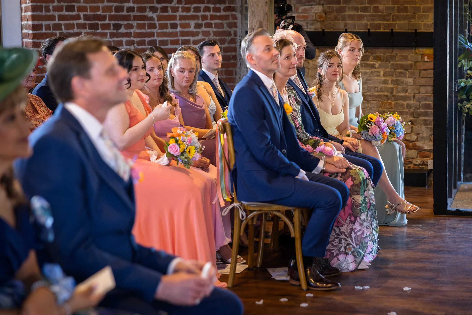 Wedding guests seated on wooden chairs inside a rustic barn with exposed brick walls watch the ceremony unfold.