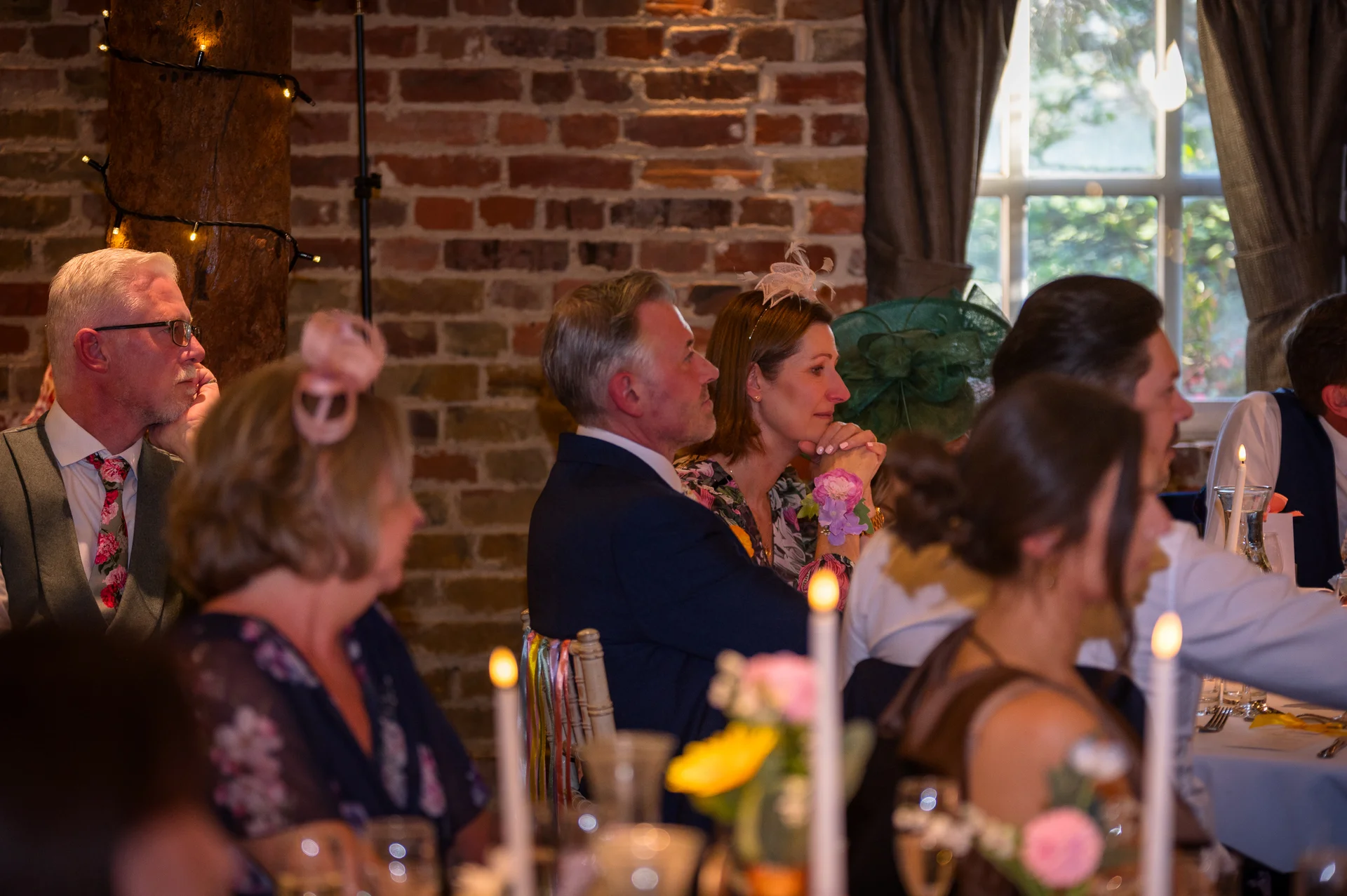 Wedding guests seated at banquet tables listening attentively during speeches in a rustic barn venue with exposed brick walls.