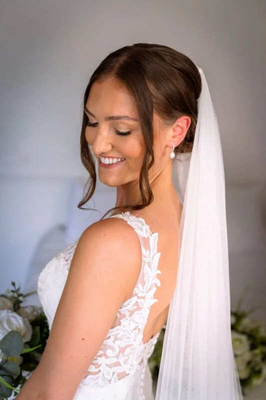 A smiling bride wearing an intricate lace gown and a simple veil looks down, holding a bouquet of white roses during her morning preparations.