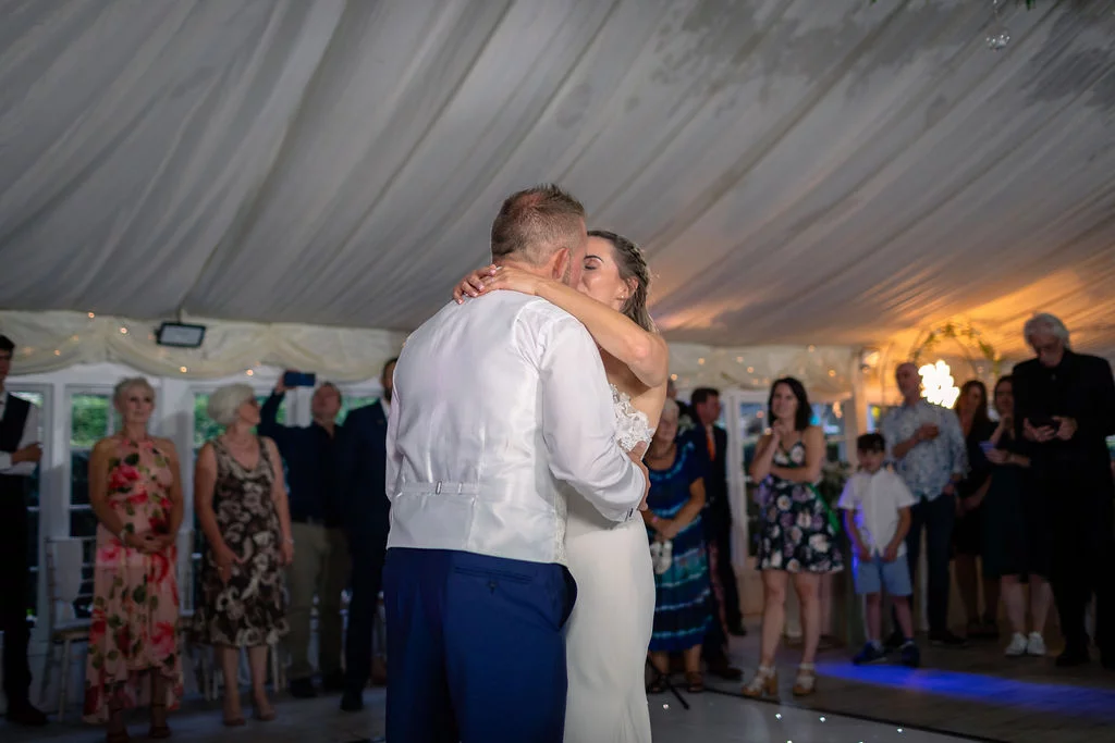 Newlyweds share an intimate embrace during their first dance inside a draped marquee surrounded by watching guests.