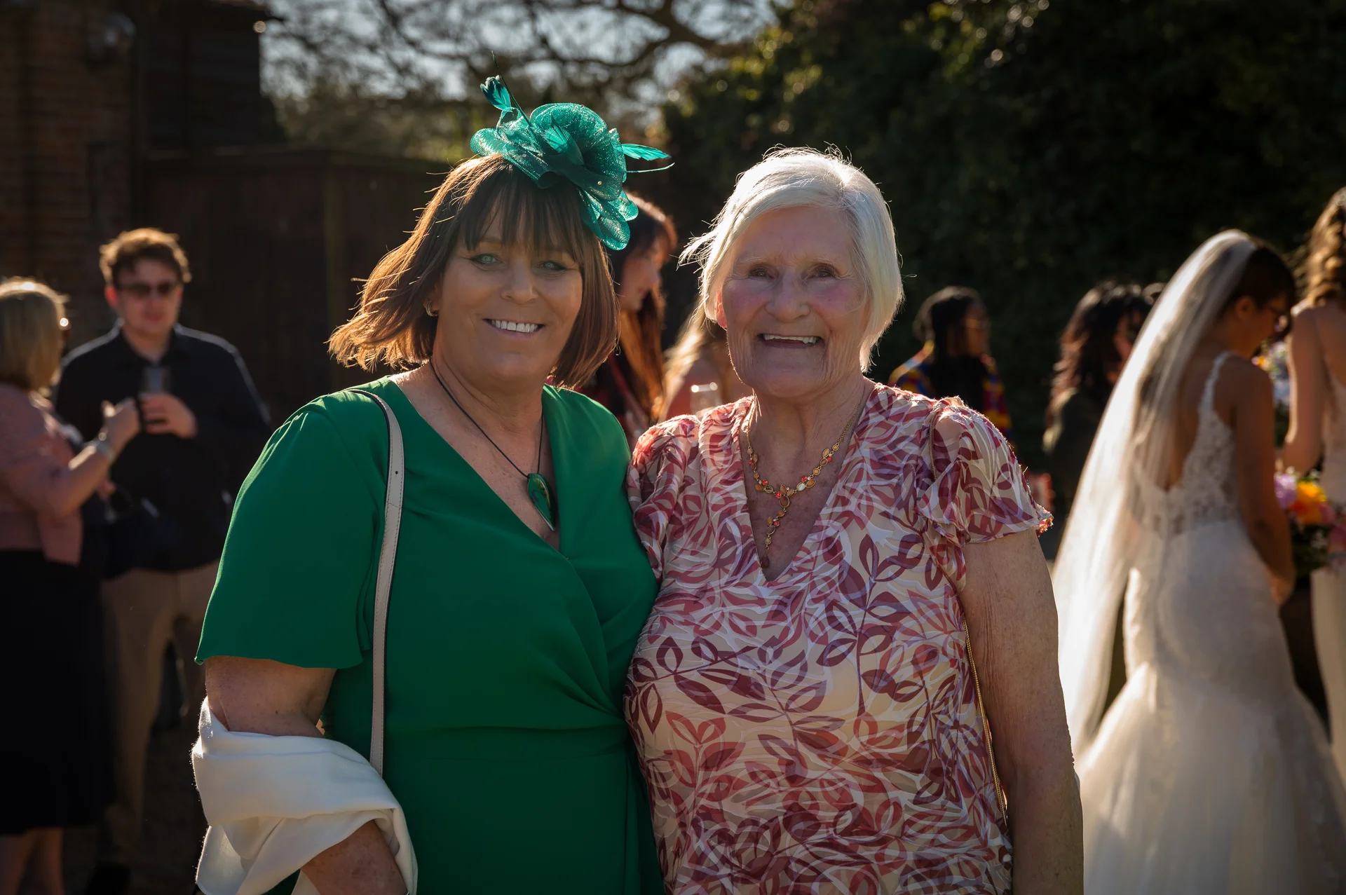 Two smiling female wedding guests stand together outdoors in the bright sunlight, with one wearing a green dress and matching fascinator.