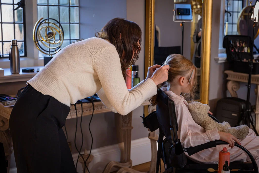 A stylist carefully styles the blonde hair of a young flower girl who sits in a chair holding a stuffed toy during morning bridal preparations near a large window.