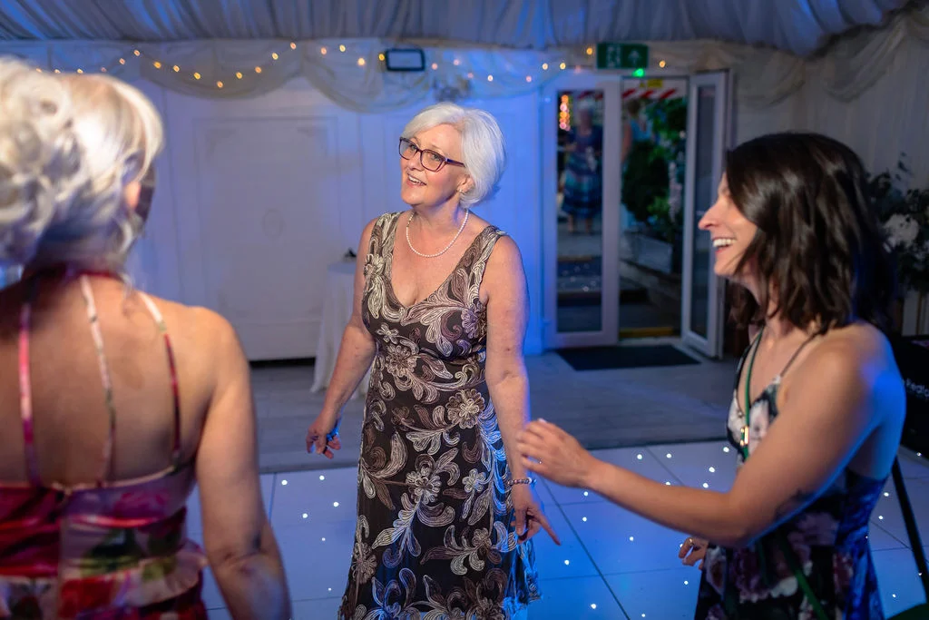 A mature woman smiles happily as she dances on an illuminated dance floor inside a marquee during a wedding evening reception.