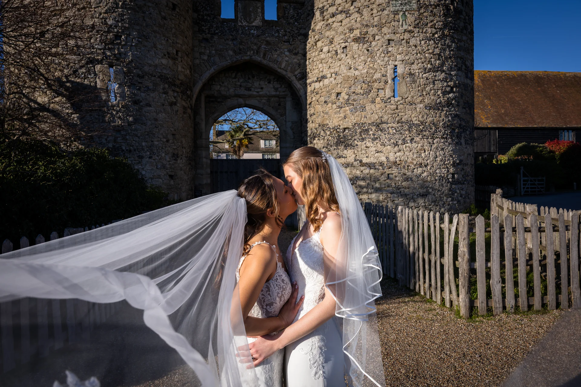 Two brides share a tender kiss in front of an ancient stone castle gateway as a translucent veil billows dramatically in the wind.