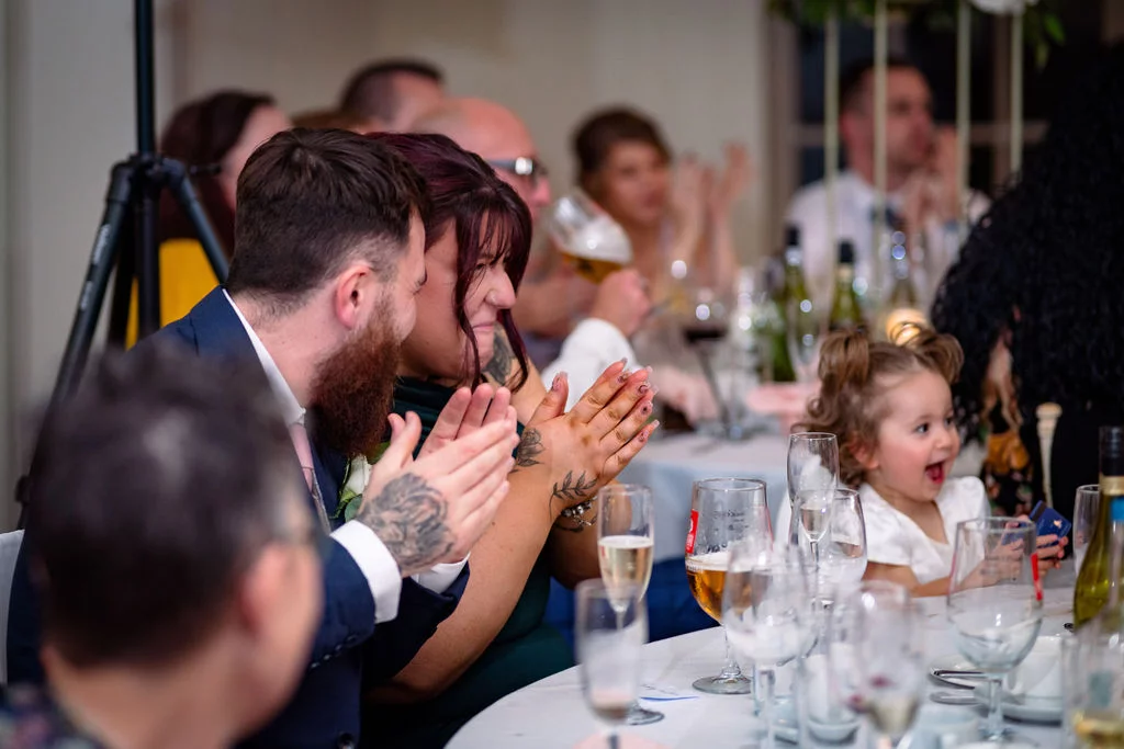 A smiling couple and guests sit at a wedding reception table, clapping and laughing during the speeches.