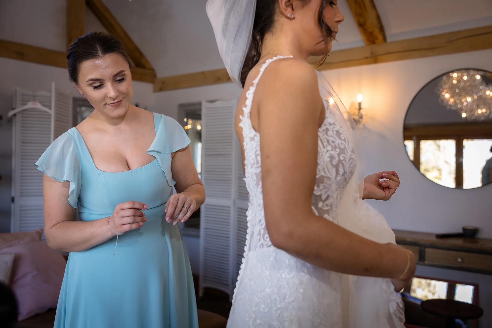 A smiling bridesmaid in a blue dress assists the bride with the final adjustments to her lace wedding gown during morning preparations.
