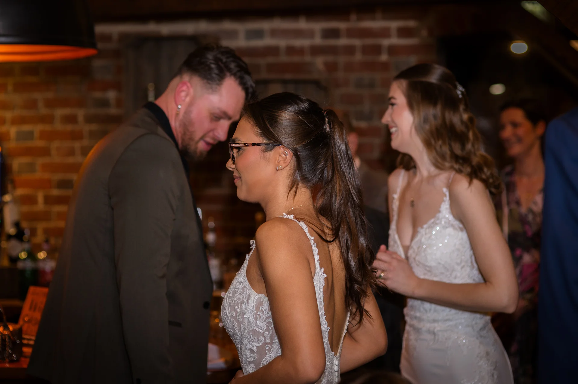 Two smiling brides in lace wedding dresses mingle with guests inside a rustic venue featuring exposed brick walls.