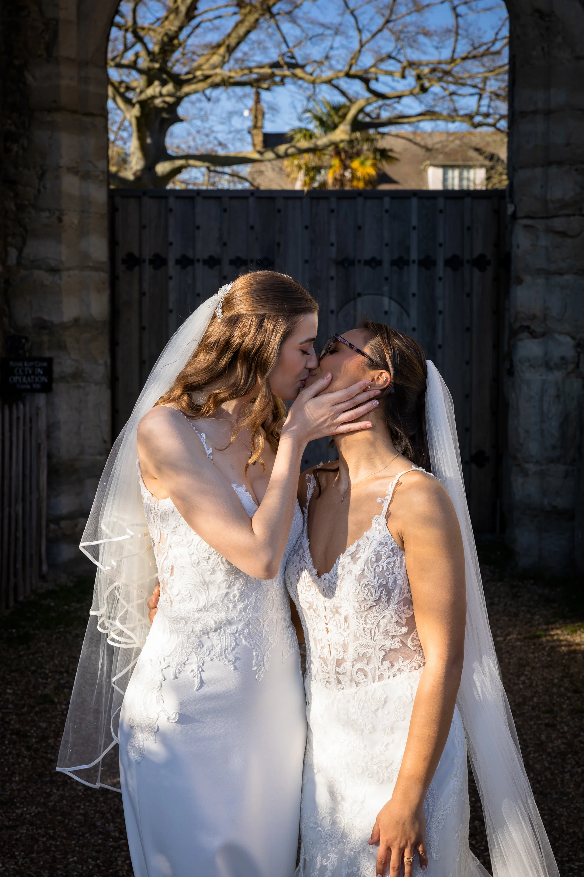 Two brides share a tender kiss beneath a rustic stone archway, both wearing lace wedding gowns and veils in the warm sunlight.