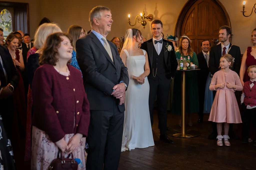 A smiling father of the bride stands proudly watching the ceremony while guests and wedding party members look on attentively inside a historic venue.