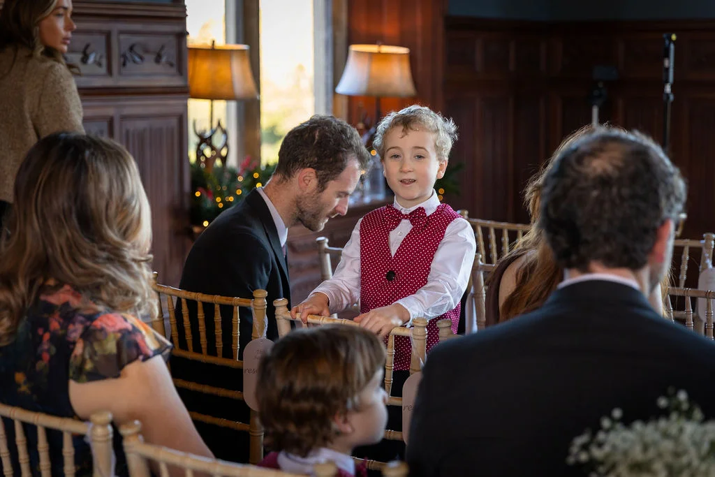 A young page boy in a red polka-dot waistcoat looks towards the camera while attending a wedding inside a wood-panelled room.
