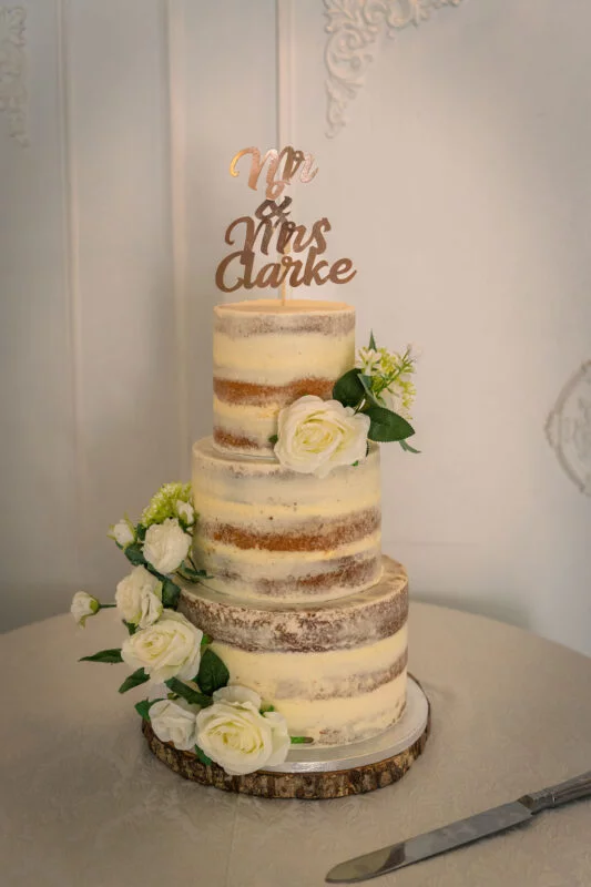 A three-tier semi-naked wedding cake adorned with white roses and a custom rose gold Mr & Mrs cake topper sits on a wooden slice stand.